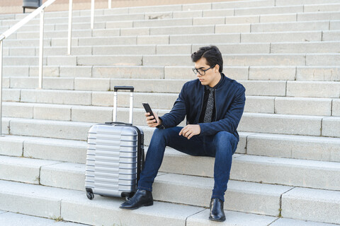 Modischer junger Geschäftsmann mit Rollkoffer, der auf einer Treppe sitzt und auf sein Mobiltelefon schaut, lizenzfreies Stockfoto