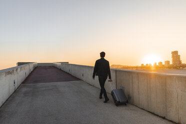 Spain, Barcelona, back view of young businessman with rolling suitcase at sunset - AFVF02342