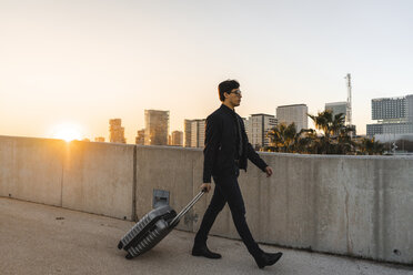 Spain, Barcelona, young businessman with rolling suitcase at sunset - AFVF02343
