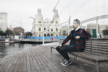 Junger Mann in Freizeitkleidung sitzt auf einer Bank am Hafen - JRFF02549