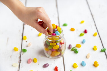 Glass of colourful sweet jellybeans on white wood, hand taking one - SARF04081