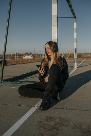 Junge Frau mit Kopfhörern und Mobiltelefon, die sich auf einem Sportplatz an ein Tor lehnt, lizenzfreies Stockfoto