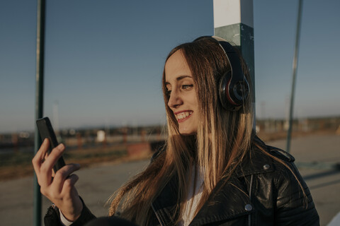 Lächelnde junge Frau mit Kopfhörern und Handy, die sich an ein Tor auf einem Sportplatz lehnt, lizenzfreies Stockfoto