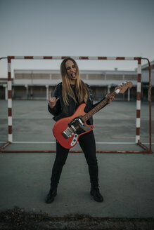 Porträt einer jungen Frau mit Gitarre auf einem Sportplatz, die ein Hupzeichen macht - DMGF00050