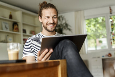 Junger Mann mit Brötchen sitzt zu Hause und benutzt ein digitales Tablet - PESF01140