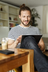 Junger Mann mit Brötchen sitzt zu Hause und benutzt ein digitales Tablet - PESF01143