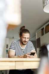 Junger Mann mit Brötchen sitzt zu Hause und benutzt sein Smartphone - PESF01151