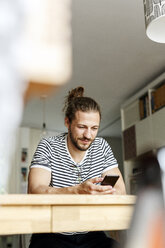 Young man with a bun sitting at home, using smartphone - PESF01151