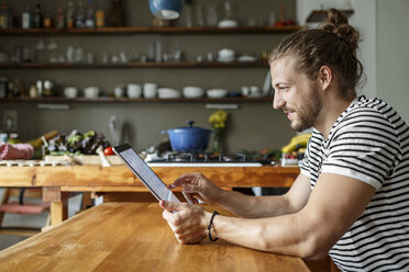 Junger Mann mit Brötchen sitzt zu Hause und benutzt ein digitales Tablet - PESF01154