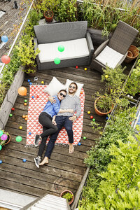 Young couple relaxing on their balcony, lying on blanket with arms around - PESF01172