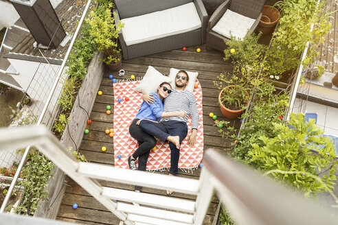 Young couple relaxing on their balcony, lying on blanket with arms around - PESF01173