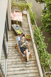 Young woman sitting in deck chair, relaxing on her balcony - PESF01186