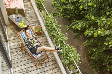 Junge Frau sitzt im Liegestuhl und entspannt sich auf ihrem Balkon - PESF01187