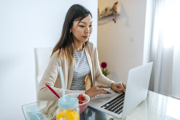 Junge Frau frühstückt, während sie am Laptop arbeitet - KIJF02262