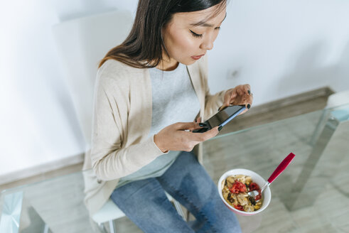 Junge Frau isst Müsli zum Frühstück, während sie ihr Smartphone benutzt - KIJF02271