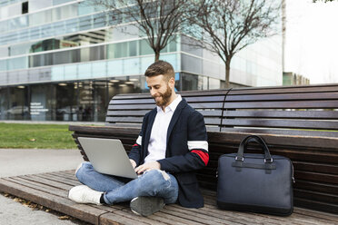 Stylish businessman sitting on bench in the city using laptop - JRFF02596