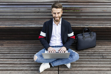 Porträt eines stilvollen Geschäftsmannes, der auf einer Bank sitzt und einen Laptop benutzt - JRFF02598