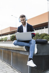 Stylish businessman sitting in the city listening to music and using laptop - JRFF02604