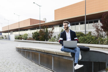 Stylish businessman sitting in the city listening to music and using laptop - JRFF02605