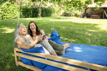 Two laughing women relaxing on a hanging bed in garden using tablet - PESF01269