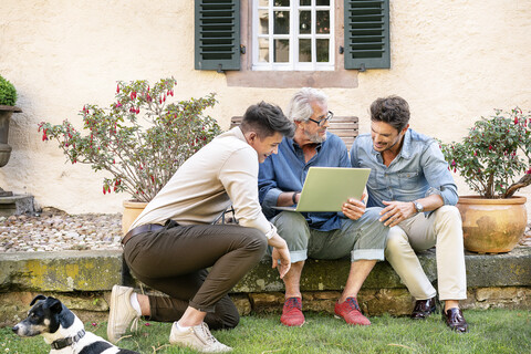 Drei glückliche Männer unterschiedlichen Alters benutzen einen Laptop im Garten, lizenzfreies Stockfoto