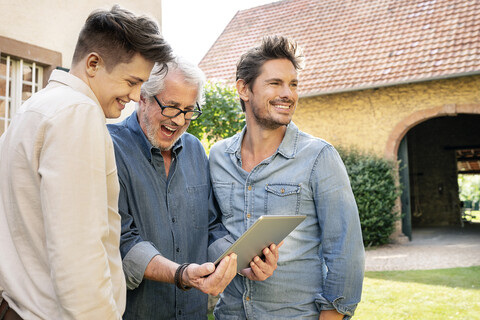 Drei glückliche Männer unterschiedlichen Alters benutzen ein Tablet im Garten, lizenzfreies Stockfoto