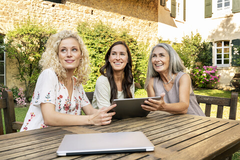 Drei Frauen unterschiedlichen Alters sitzen am Gartentisch und benutzen ein Tablet, lizenzfreies Stockfoto