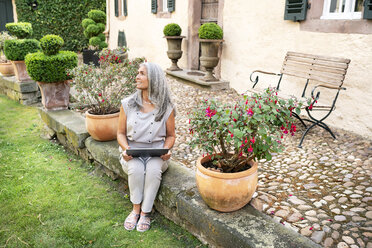 Frau mit langen grauen Haaren sitzt auf der Terrasse eines Landhauses und benutzt ein Tablet - PESF01350