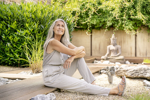 Lächelnde Frau mit langen grauen Haaren sitzt auf der Terrasse im Garten - PESF01360