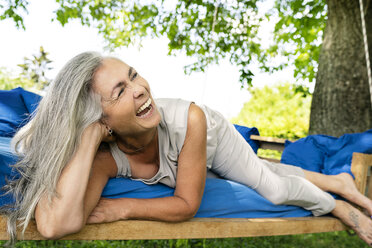 Lachende Frau mit langen grauen Haaren auf einem Bett im Garten liegend - PESF01368