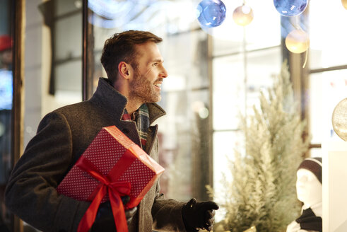 Lächelnder Mann mit Weihnachtsgeschenk, der in ein Schaufenster schaut - ABIF01157