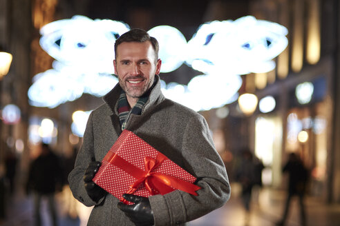 Porträt eines lächelnden Mannes mit Weihnachtsgeschenk in der Fußgängerzone am Abend - ABIF01159