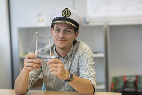 Portrait of smiling businessman sitting at desk in office wearing captain's hat and holding model ship - PAF01905