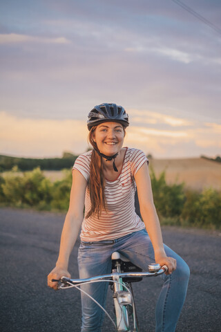 Porträt einer lächelnden jungen Frau mit Fahrrad auf einer Landstraße in der Abenddämmerung, lizenzfreies Stockfoto