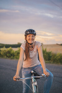 Porträt einer lächelnden jungen Frau mit Fahrrad auf einer Landstraße in der Abenddämmerung - JSCF00156