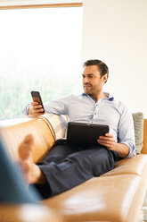 Reifer Mann schaut auf sein Smartphone, während er sich auf dem Sofa zurücklehnt - CUF48514
