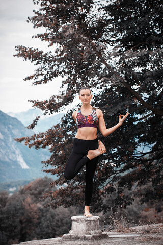 Frau übt Yoga, balanciert auf einem Bein auf einem Sockel, Berglandschaft, Domodossola, Piemonte, Italien, lizenzfreies Stockfoto