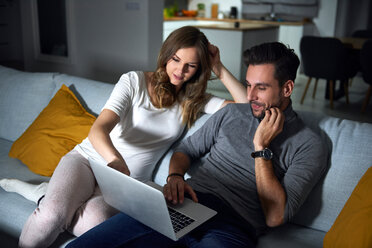 Young couple sitting on sofa in evening looking at laptop - CUF49107