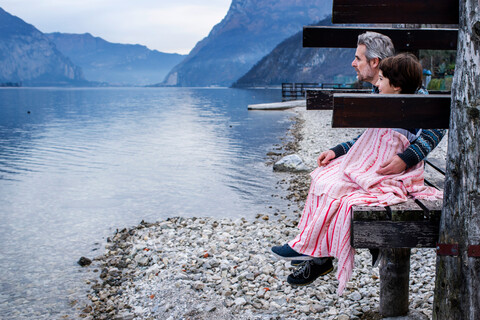 Junge und Vater in eine Decke eingewickelt auf einem Steg am See, Seitenansicht, Comer See, Onno, Lombardei, Italien, lizenzfreies Stockfoto