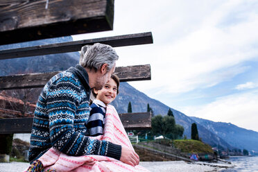 Boy and father wrapped in blanket on lakeside pier, side view, Lake Como, Onno, Lombardy, Italy - CUF49230