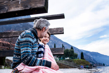 Junge und Vater in eine Decke eingewickelt auf einem Steg am See, Seitenansicht, Comer See, Onno, Lombardei, Italien - CUF49230