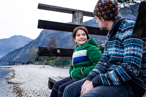 Junge und Vater sitzen plaudernd am Seeufer, Comer See, Onno, Lombardei, Italien, lizenzfreies Stockfoto