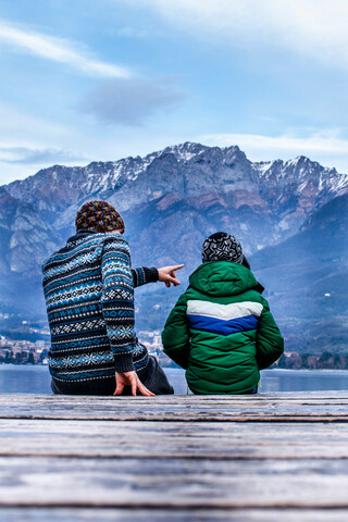 Boy and father sitting pointing on lakeside pier, rear view, Lake Como, Onno, Lombardy, Italy stock photo