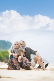 Portrait of happy senior hippie couple sitting side by side on the beach - SBOF01703