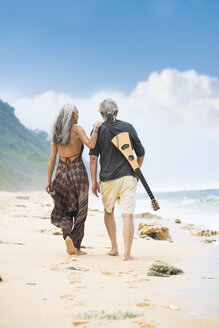 Rückenansicht eines älteren Hippie-Paares mit Gitarre, das Seite an Seite am Strand spazieren geht - SBOF01711