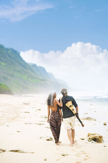 Rückenansicht eines älteren Hippie-Paares mit Gitarre, das Seite an Seite am Strand spazieren geht - SBOF01712
