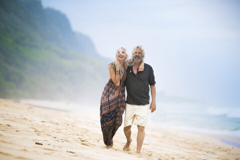 Glückliches älteres Hippie-Paar, das Seite an Seite am Strand spazieren geht, lizenzfreies Stockfoto