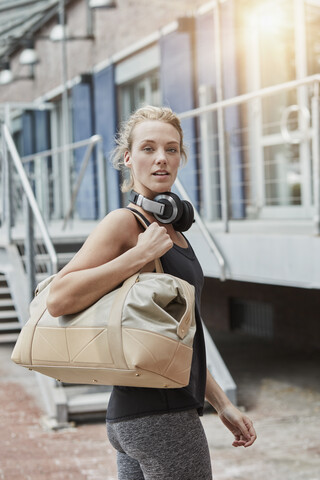 Porträt einer blonden jungen Frau mit Kopfhörern und Sporttasche vor einem Fitnessstudio, lizenzfreies Stockfoto