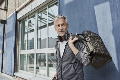 Porträt eines reifen Mannes mit Kopfhörern und einer Sporttasche in Tarnfarben vor einem Fitnessstudio - RORF01729