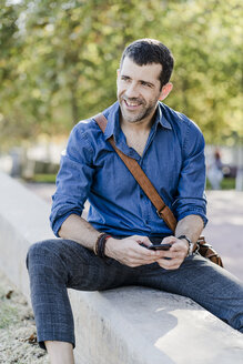 Porträt eines lächelnden Mannes mit Smartphone, der an einer Wand sitzt - GIOF05705
